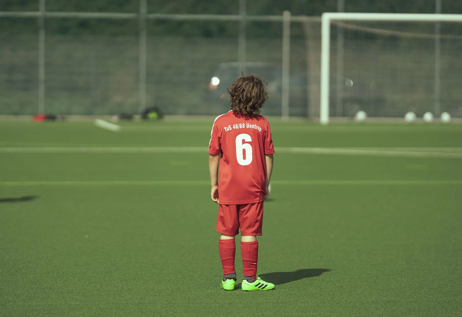 A boy seen from behind in a red jersey numbered six, standing alone on a green artificial turf field with a goal in the distance.