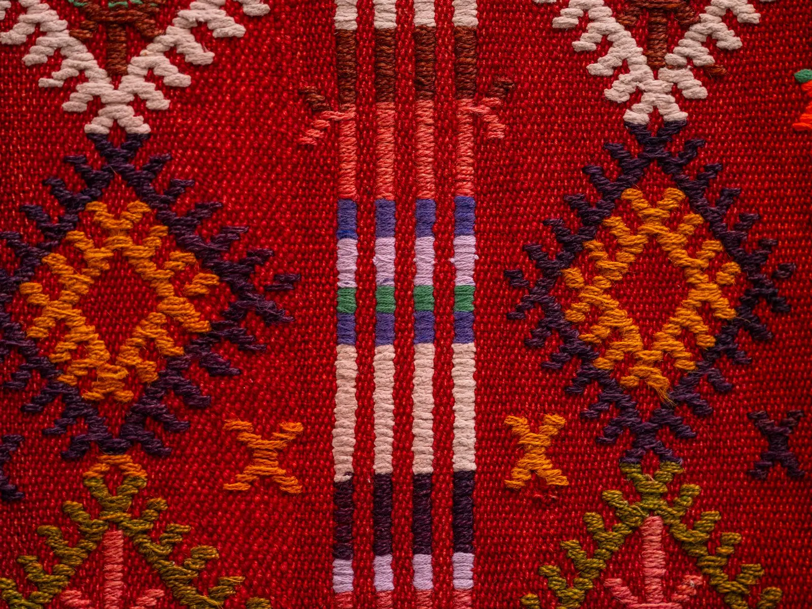 A close-up of a red kilim rug woven with orange diamonds, dark purple crosses, and a narrow striped band of blue, green, and cream.