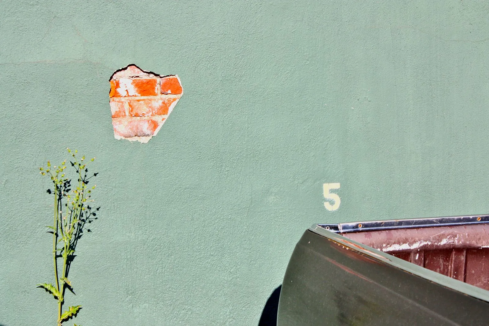 A sage-green wall with a ragged patch of plaster missing, revealing red brick underneath, beside a stencilled house number and the dark hull of an upturned boat.
